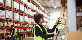 Female warehouse worker stacking boxes onto the rack. Woman working in large distribution warehouse. she is wearing uniform and reflective clothing, with colleagues working in background.